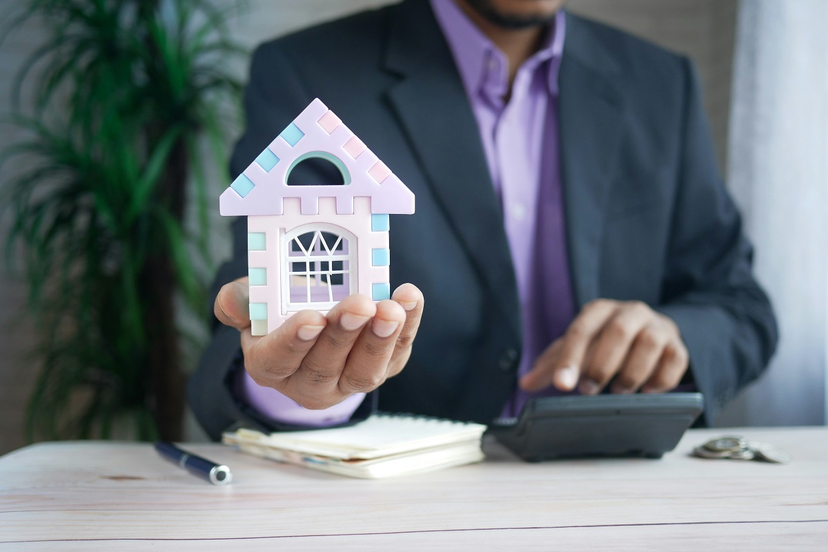man in a suit holding a miniature house while using a calculator